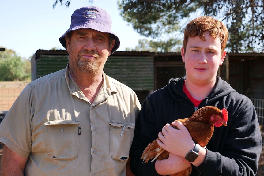A father in a blue bucket hat and brown shirt stands next to his son who's wearing a blue jumper and holding a brown chicken.
