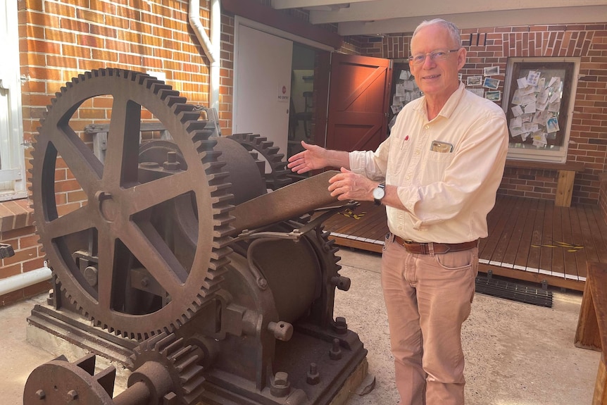 Man stands next to a large rusted sugar cane crusher, with a mechanical wheel and sloped metal feeder. 