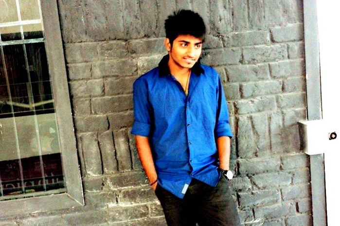 A young man in a blue shirt and dark trousers leans against a bluestone wall.