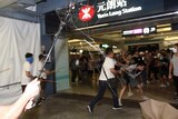 Viewing from within the scrum of a fight, you view three men in white shirts carrying poles and bamboo sticks attacking people.