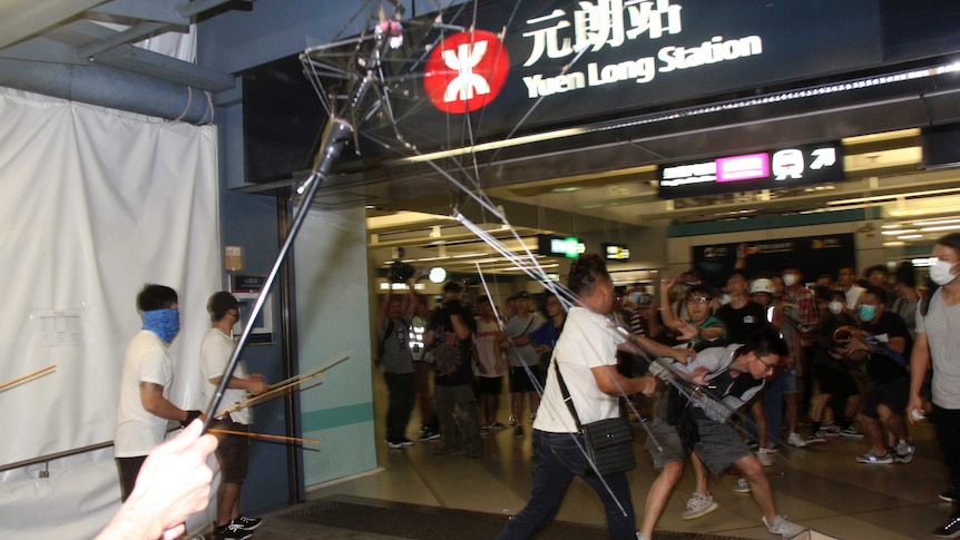 Viewing from within the scrum of a fight, you view three men in white shirts carrying poles and bamboo sticks attacking people.