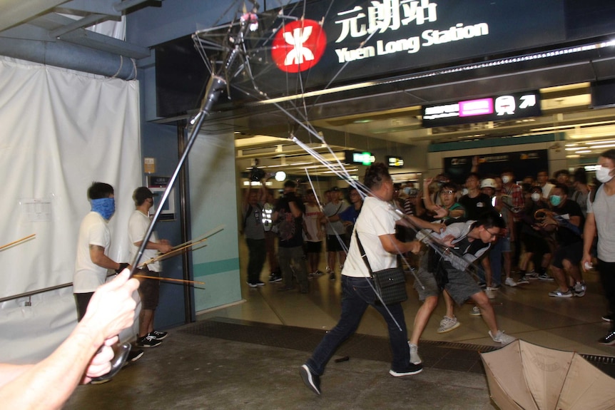 Viewing from within the scrum of a fight, you view three men in white shirts carrying poles and bamboo sticks attacking people.