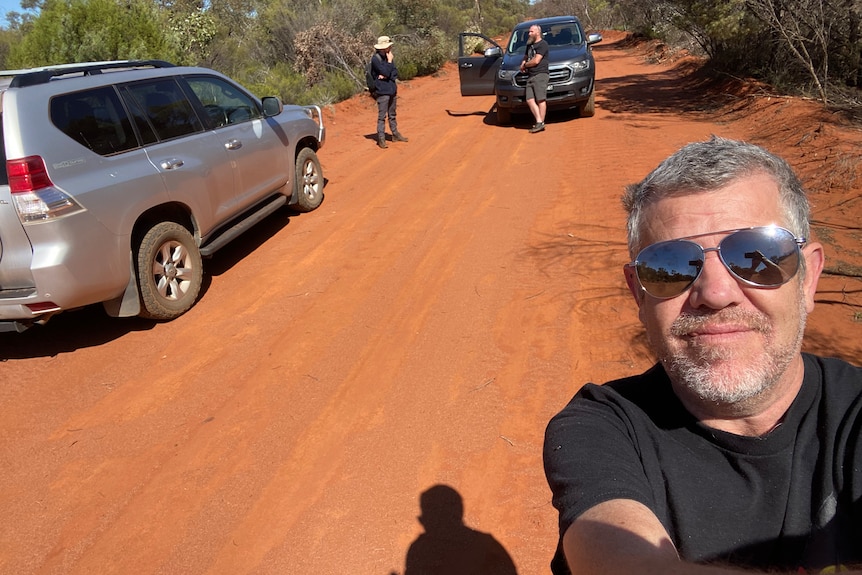A man in front of a red dirt road in bushland area, with two cars and a couple of people standing behind him.