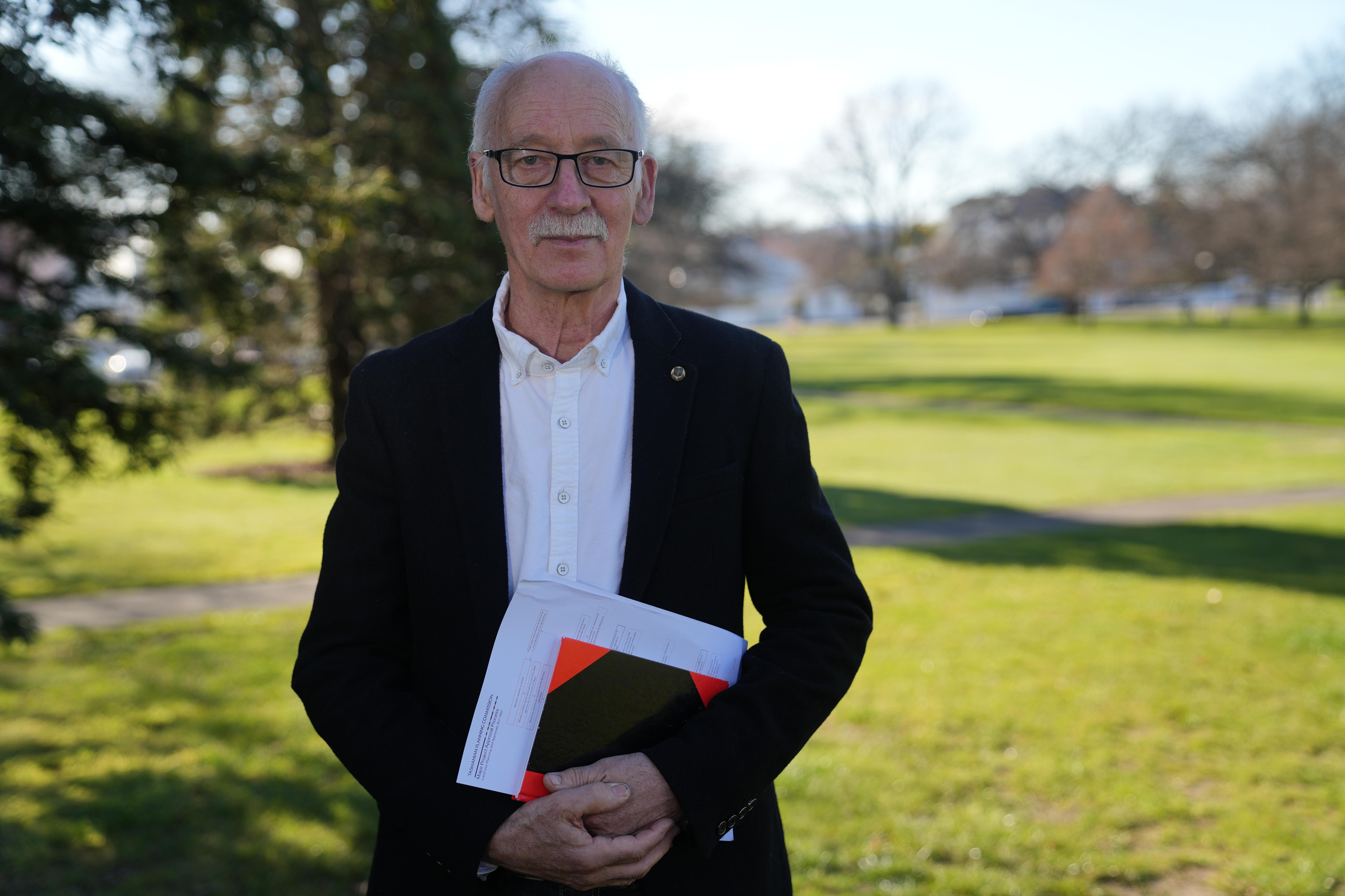 He stands in a park wearing formal clothes. He has white hair. He holds a notebook and looks at the camera.