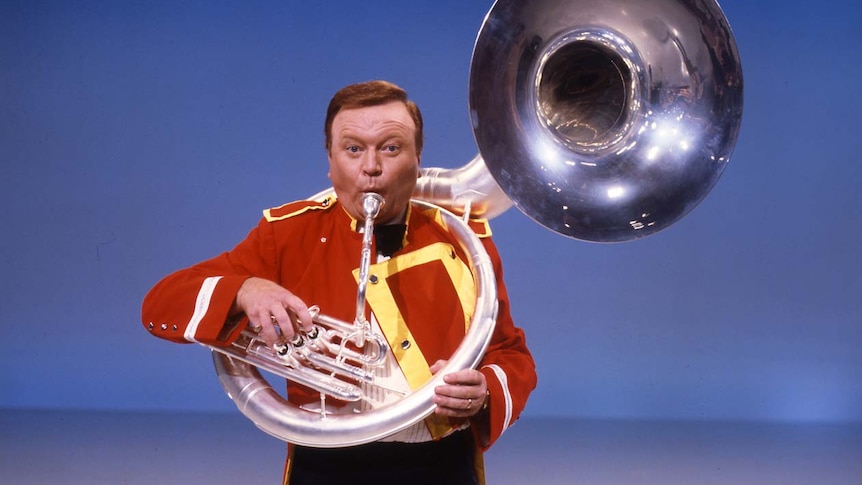 Bert Newton dress in a band uniform, blowing through a tuba.