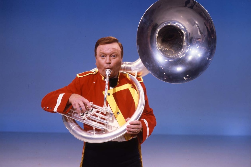 Bert Newton dress in a band uniform, blowing through a tuba.