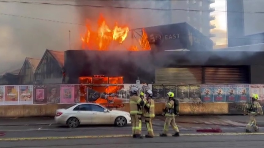 Firefighters stand on the street as orange flames engulf a building on a boarded-up site.