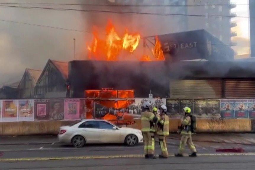 Firefighters stand on the street as orange flames engulf a building on a boarded-up site.