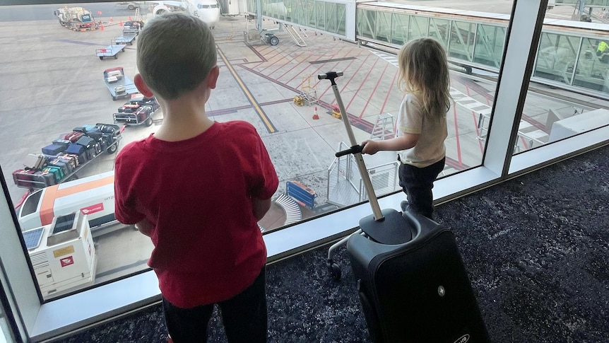 Two children stare out of an airport terminal towards the tarmac, one holding a suitcase by the handle.