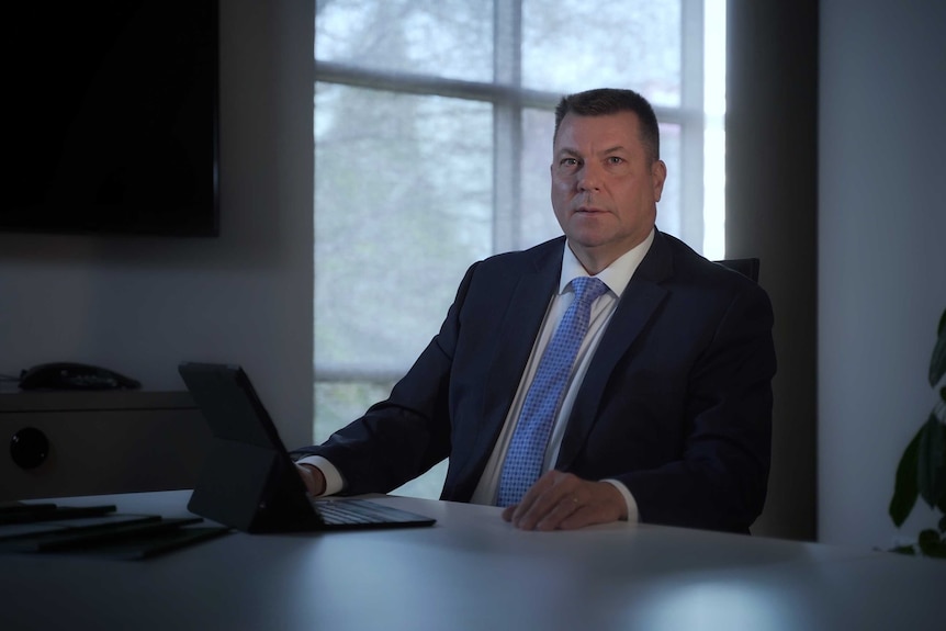 Marc White, wearing a suit and looking at the camera, sits in front of a laptop in an office.