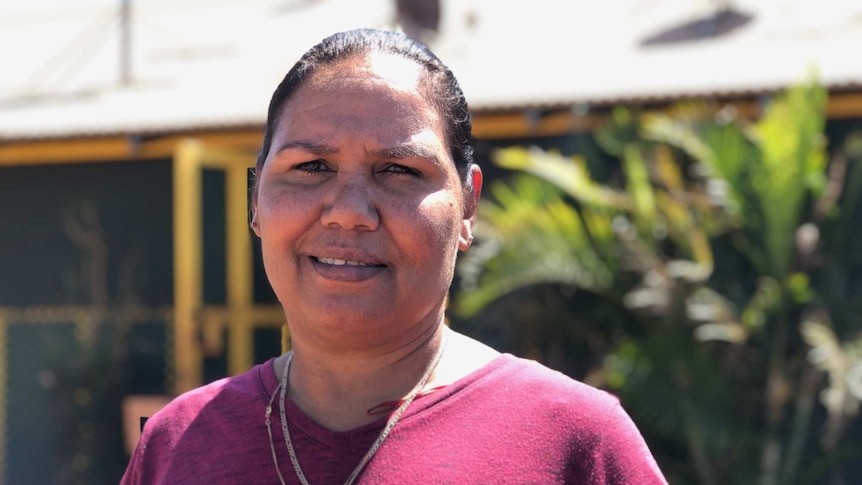 Patricia Riley, an indigenous woman, stands in front of her house, which has satellite dishes, she is wearing a burgundy t-shirt