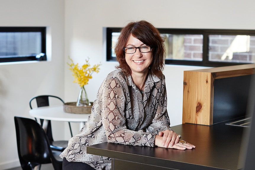 Sally Wills sits on a silver stool at a black kitchen bench in a modern white-walled house.