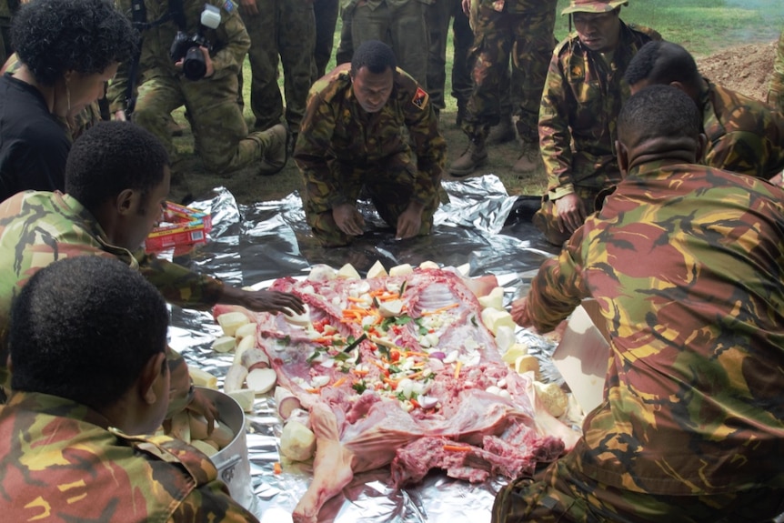 An animal carcass is splayed out on a sheet of foil with vegetables, with PNG men in army fatigues gathered around it.