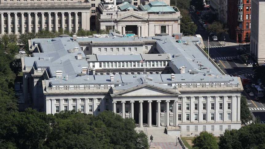 An aerial shot shows a large building with composite columns surrounded by trees and other large buildings