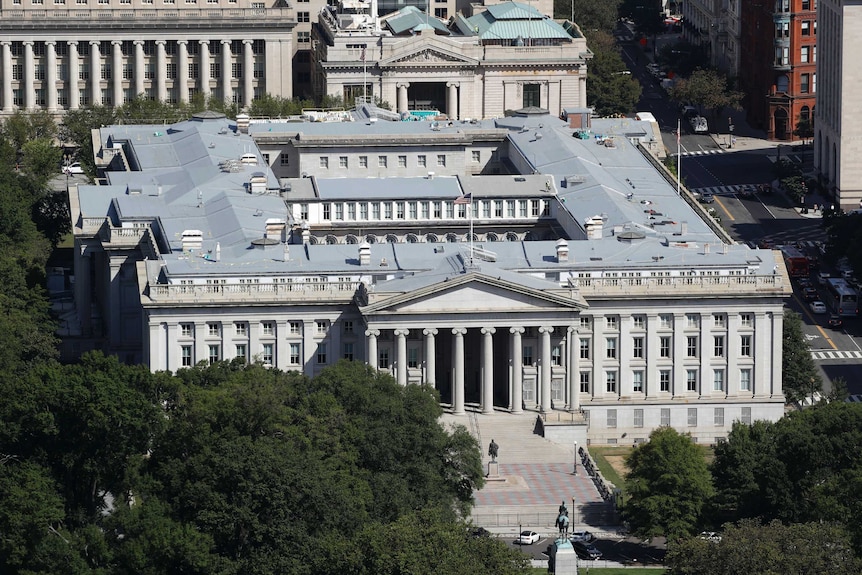 An aerial shot shows a large building with composite columns surrounded by trees and other large buildings