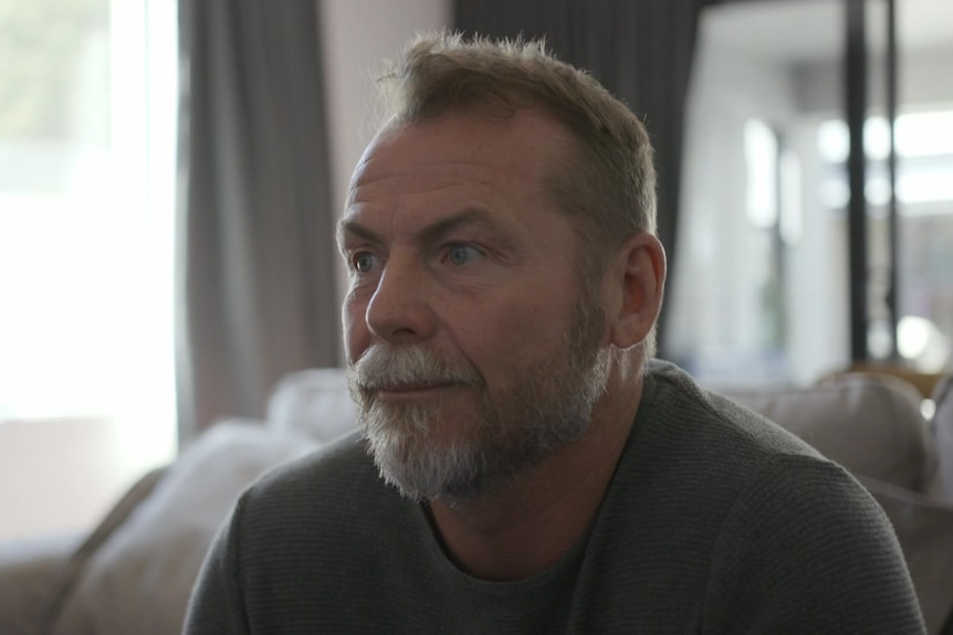 A middle-aged man with a beard sits on a sofa, looking off into the distance