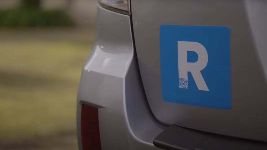 The rear bumper of a silver SUV, with a square blue plate affixed with a prominent white 'R' on it