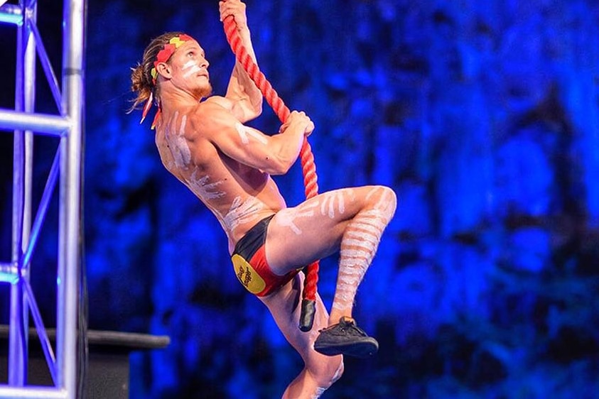 A man in Aboriginal flag colours and cultural face paint swings on a rope across an obstacle course