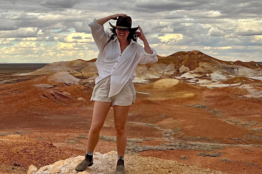 Woman wearing white holding on to her wide brimmed hat with clouds over red dirt landscape
