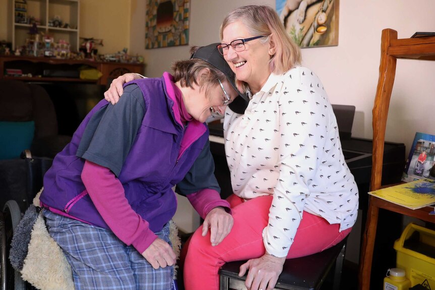 A middle-aged woman with hydrocephalus hugs her mother in their living room.