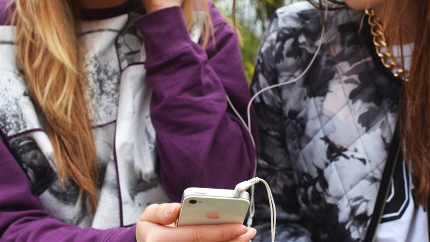 Two young women use an iPhone with headphones.