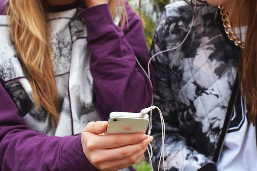 Two young women use an iPhone with headphones.