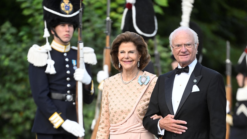 Queen Silvia dressed in a white gown and King Carl XVI Gustaf wearing a dark suit walk together in front of a guard.