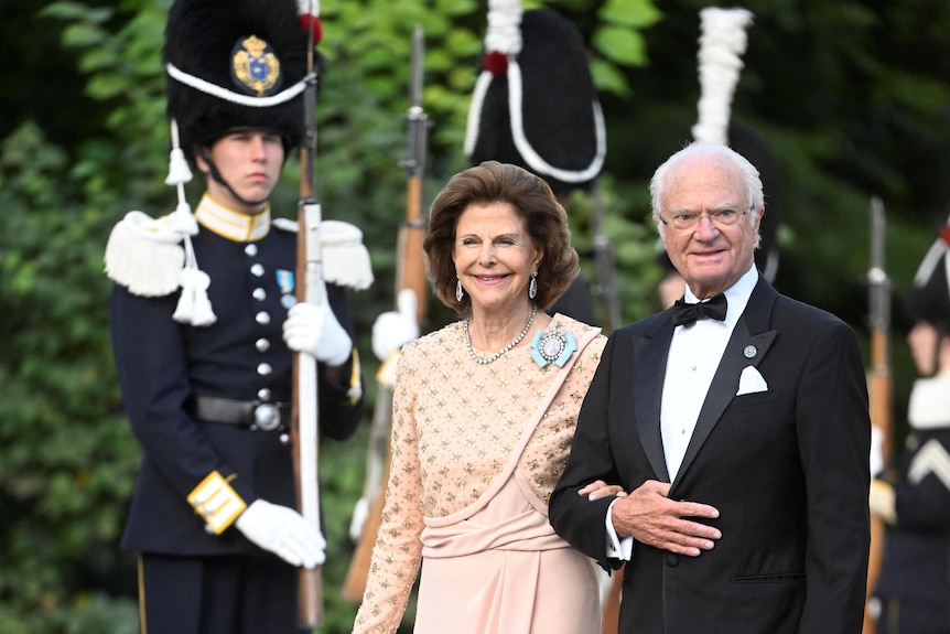 Queen Silvia dressed in a white gown and King Carl XVI Gustaf wearing a dark suit walk together in front of a guard.