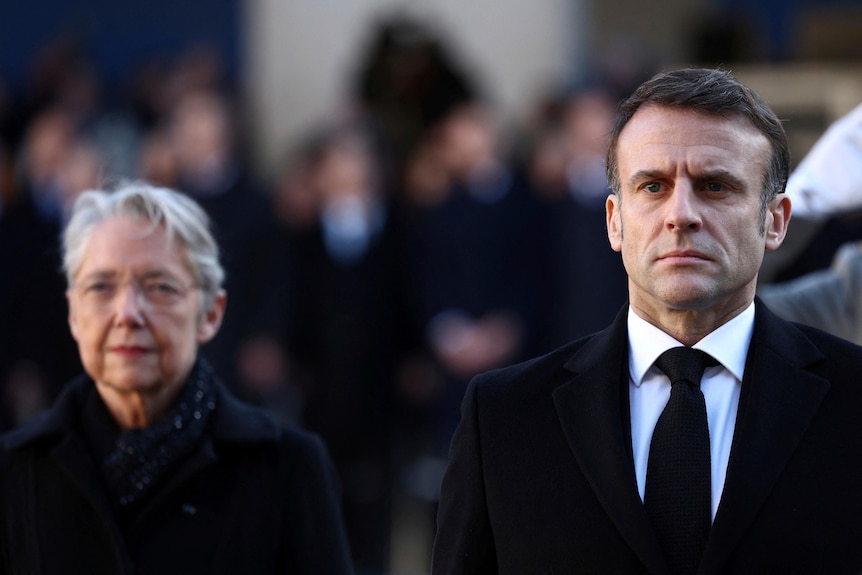 President Emmanuel Macron in a black suit stands in front of Élisabeth Borne