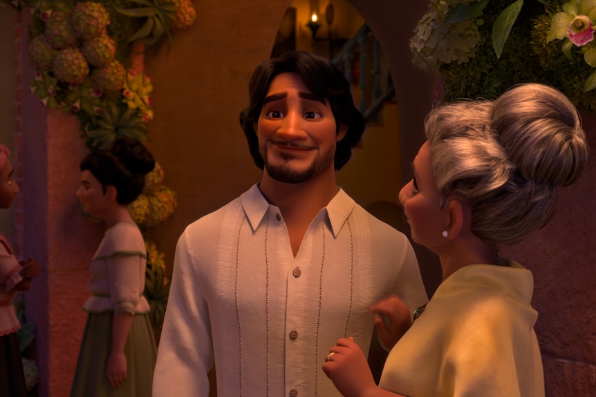 Animated, tall and bearded Latino with soft dark hair and white collared shirt chats with grey-haired woman in beige blouse.