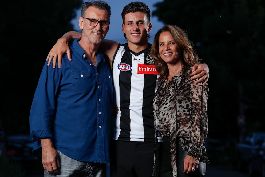 A young footballer wearing a Collingwood jersey smiles at the camera as he stands between his dad and mum.