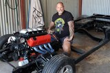 Will Barabas, a tall, stocky man with tattoos and a beard, sits in a black chassis with a red engine, inside a shed.