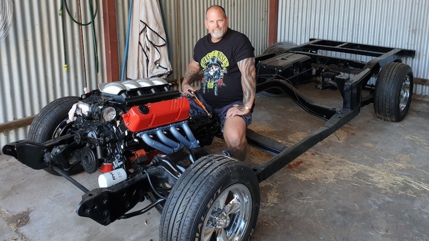 Will Barabas, a tall, stocky man with tattoos and a beard, sits in a black chassis with a red engine, inside a shed.