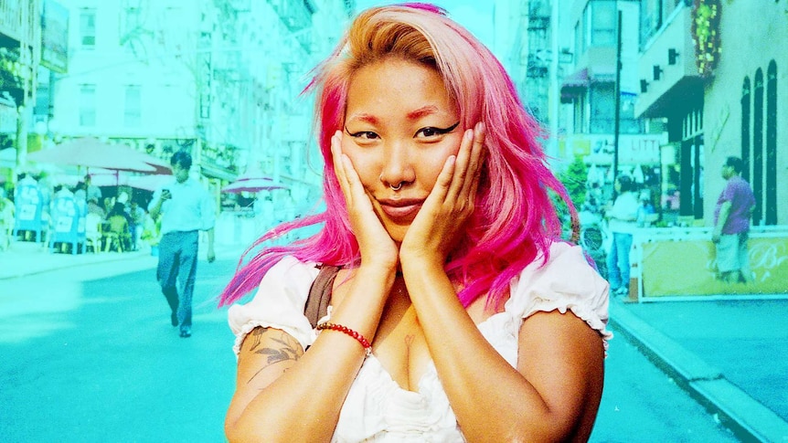 Smiling woman standing on footpath of busy street holds her hands to her cheeks for a story about Asian flush when drinking.