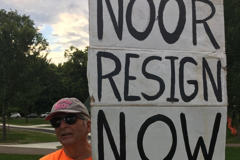 A man holds a sign reading Noor resign now.
