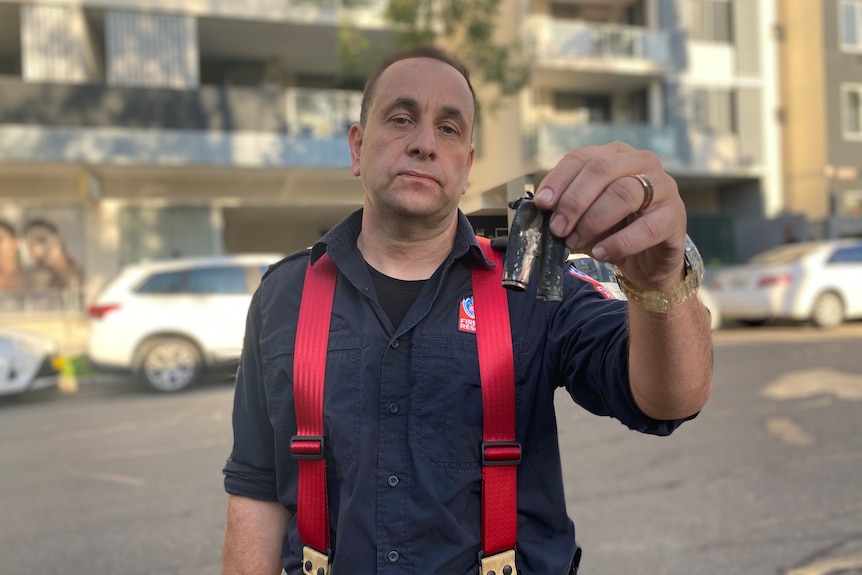 A firefighter standing on a street holds up two fried battery cells up to the camera.