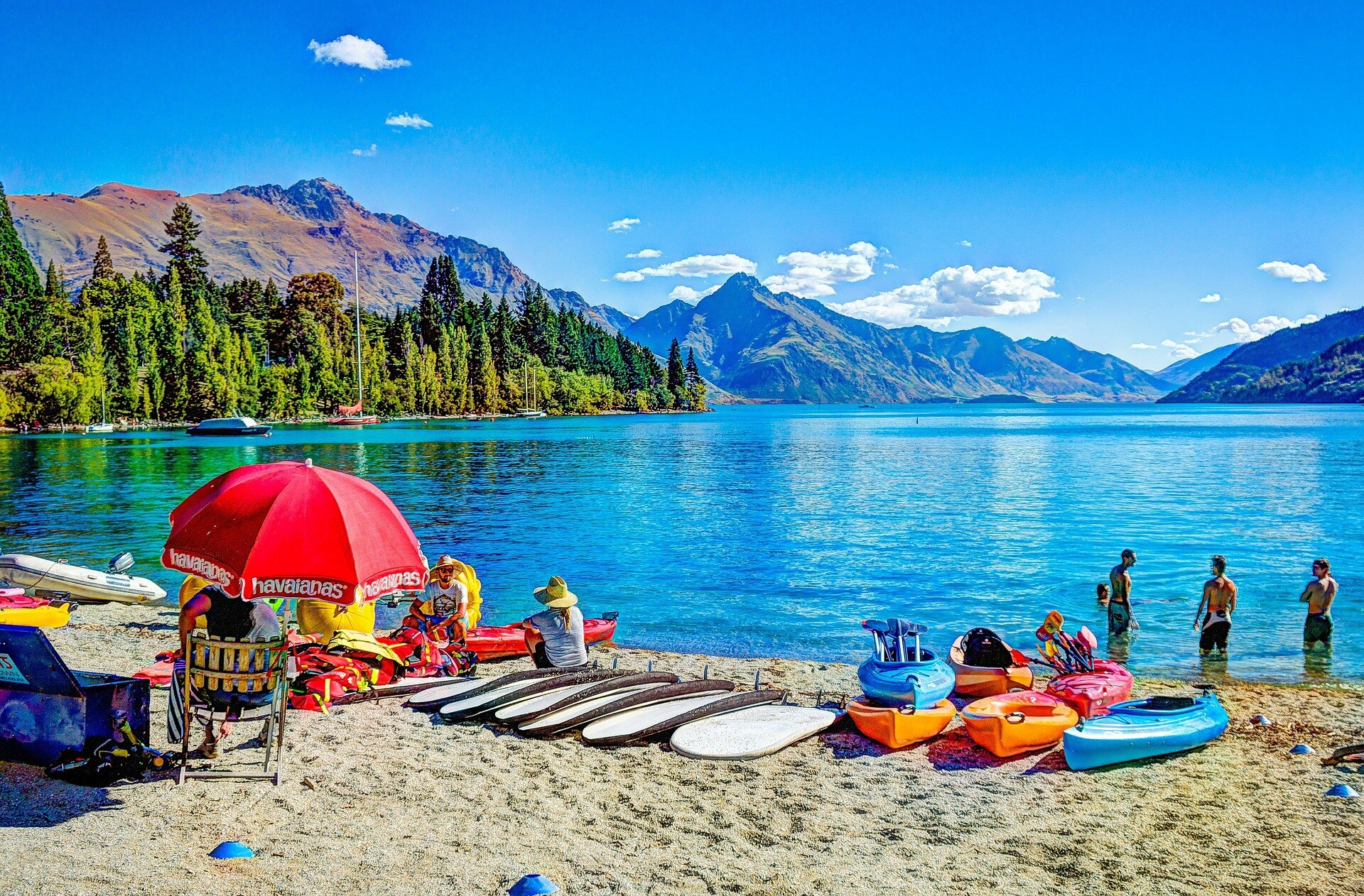 A beach umbrella, boards and people on a beach in front of a lake and mountains