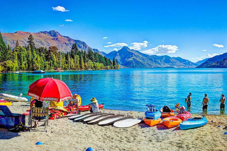 A beach umbrella, boards and people on a beach in front of a lake and mountains