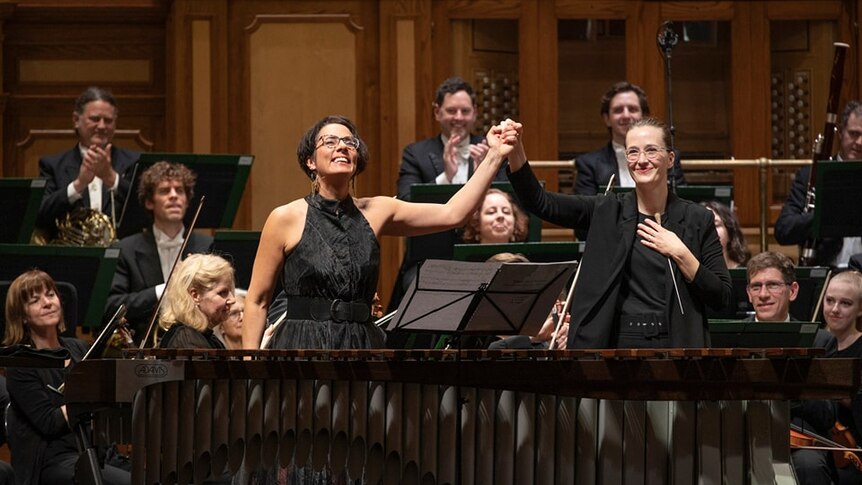 Conductor Elena Schwarz holds Claire Edwardes's hand up in the air in triumph in front of the Adelaide Symphony Orchestra.