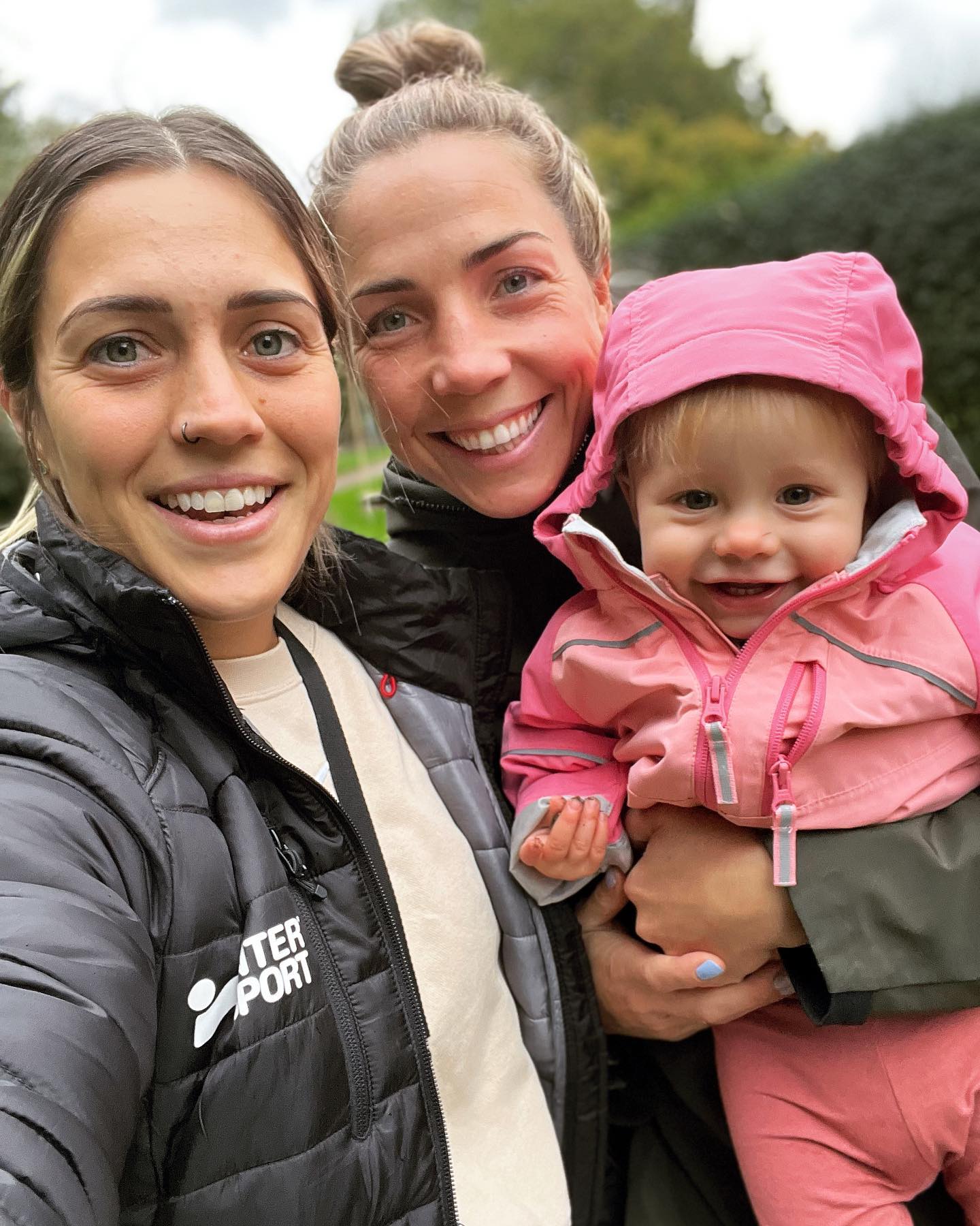 Selfie two blonde women holding a toddler girl and she wears a pink hooded jumper