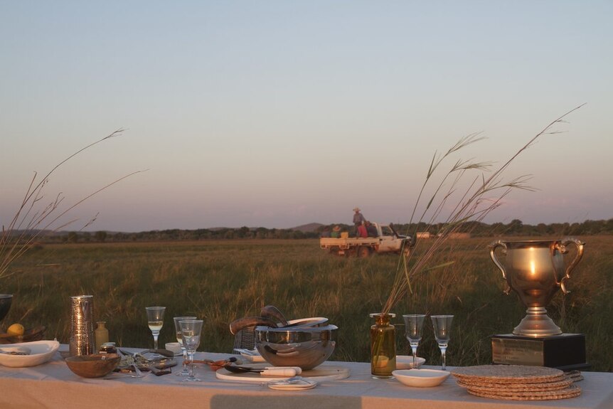 A table set with wine glasses, platters, serving bowls and other homewares in the middle of a paddock as the sun sets.