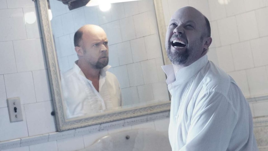 A man turns away from a mirror and laughs manically. His reflection stares at him frowning.