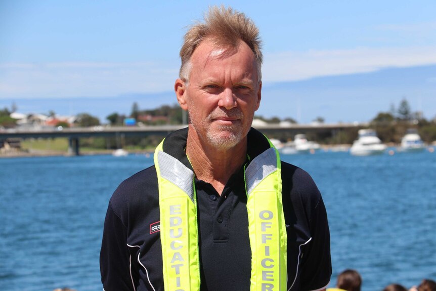 A middle-aged man wearing a life jacket stands on a jetty with the Swan River in the background
