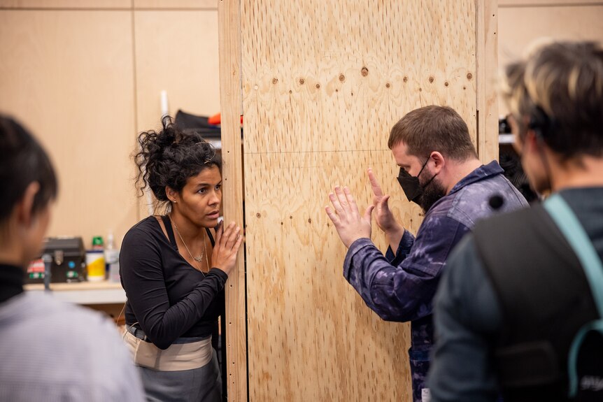 In a rehearsal room, Zahra Newman peers around a wooden set at a masked Kip Williams, giving her direction.