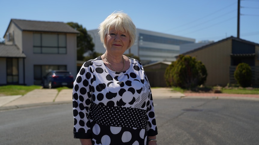 A woman with short blonde hair wearing a black and white top and necklace stands in a residential street