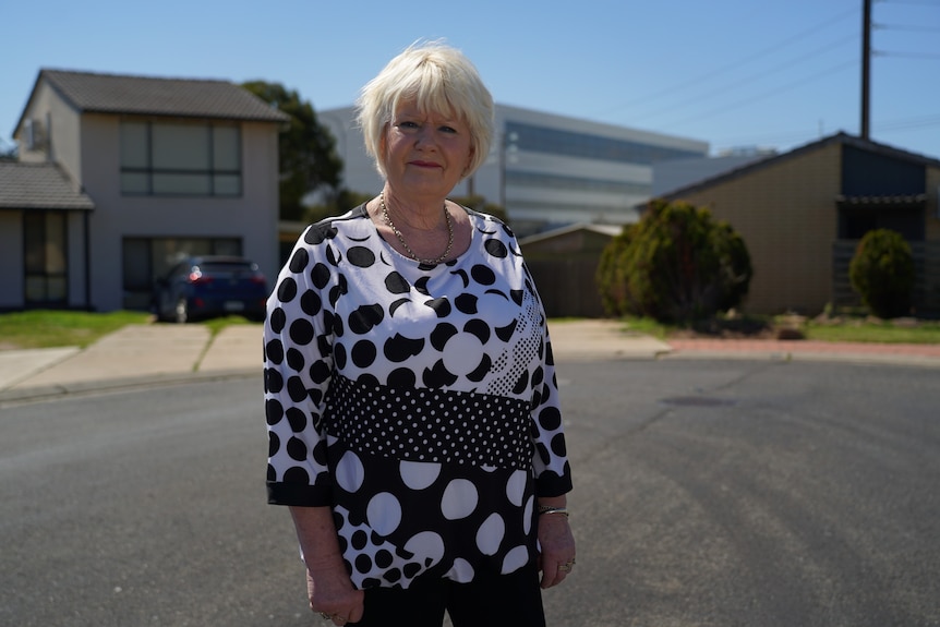 A woman with short blonde hair wearing a black and white top and necklace stands in a residential street