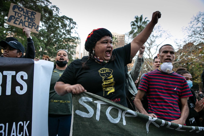 A woman wearing a hat and black lives matter shirt raises her left fist.