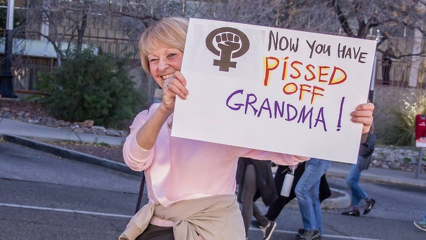 An smiling elderly woman with a bum bag holds up a sign that reads Now You've Pissed Off Grandma