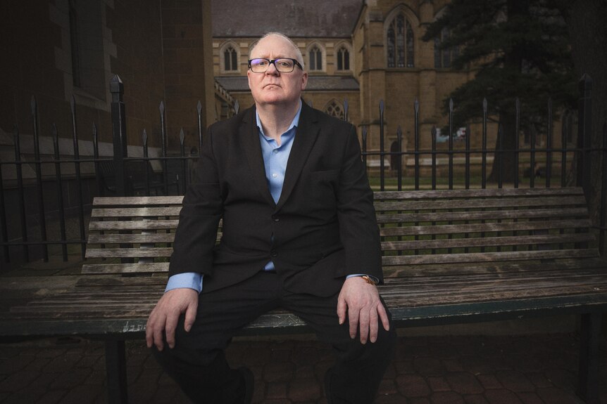 A man wearing glasses sits on a bench in front of a church