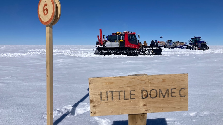 Red tractor in background on snow, a wooden sign in the foreground that says "Little Dome C"